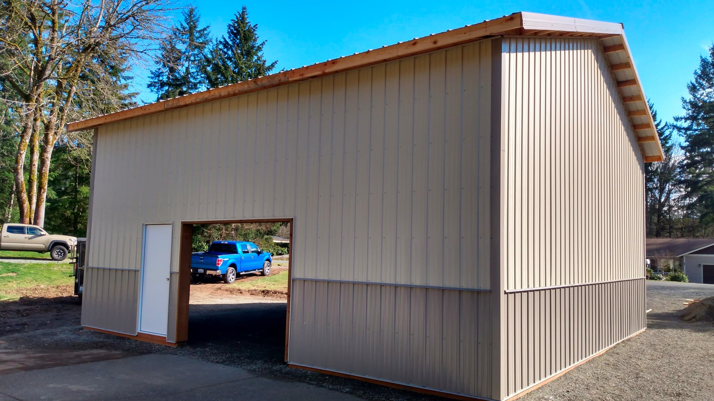 Detailed side view of a 24'x36' sloped-roof metal building constructed by Probuilt in Auburn. Features include light stone vertical metal panels, ash grey wainscoting, exposed wooden eaves with an 18-inch overhang, and a convenient side entry door. The thoughtful design provides excellent drainage while maintaining a stylish appearance for residential properties.